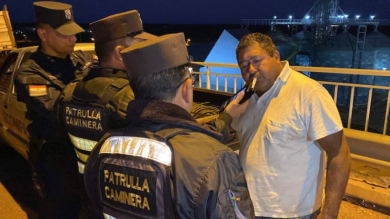 Tres agentes de Patrulla Caminera en uniforme revisan a un hombre robusto con camiseta blanca, cerca de instalaciones industriales nocturnas.