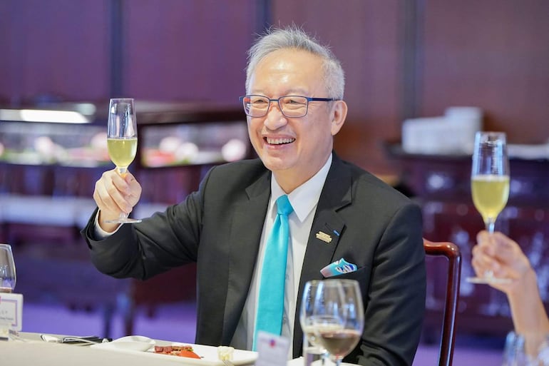 François Chih-chung Wu sonríe mientras sostiene una copa con una bebida amarilla, vestido con traje negro y corbata azul en un ambiente elegante.