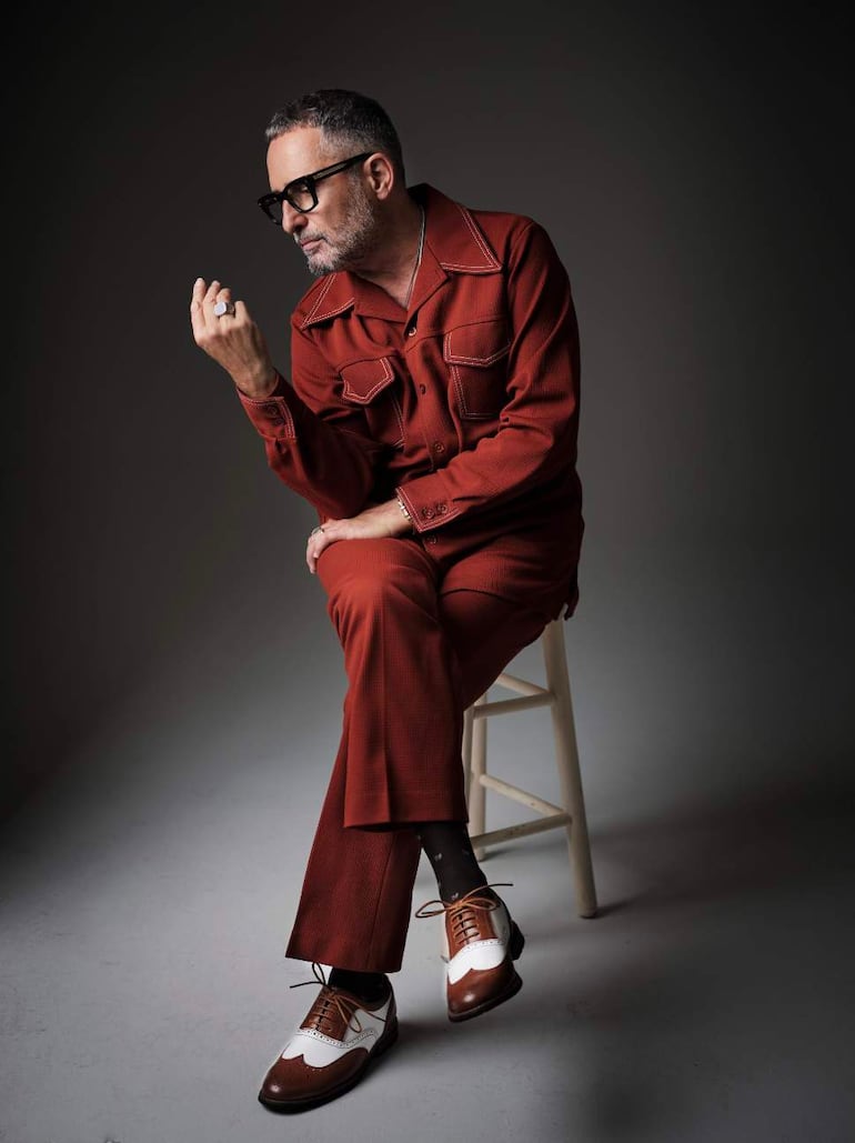 Jorge Drexler con gafas, sentado en un taburete, viste camisa y pantalones rojos, mostrando delicadamente un anillo.