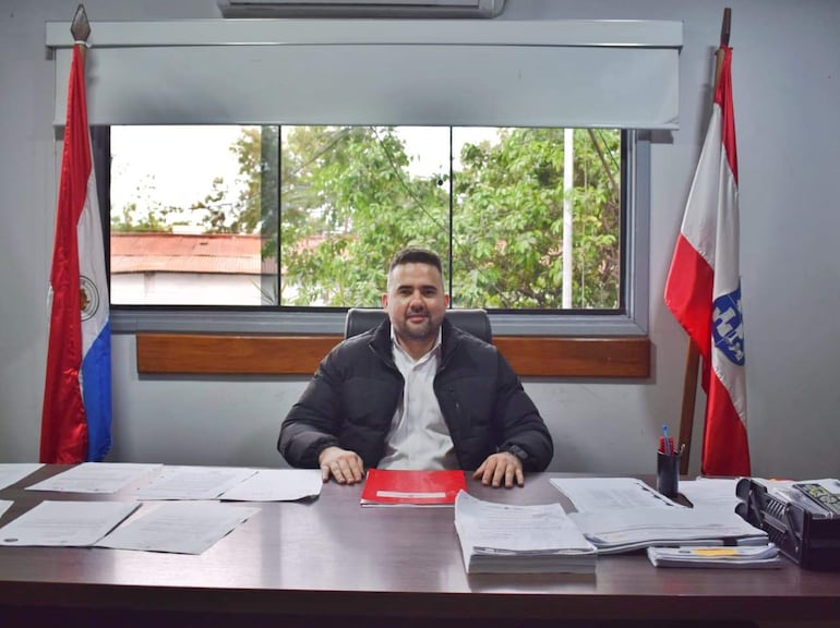 Hombre en abrigo oscuro y camisa blanca, sentado en escritorio con documentos, rodeado por banderas en un ambiente de oficina.