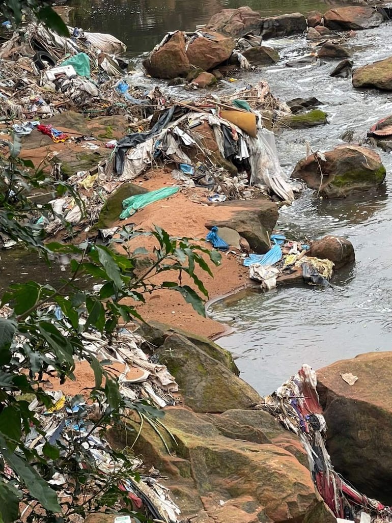 Vista del arroyo con residuos de telas y plásticos acumulados sobre rocas y arena, sin personas visibles.