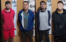 Cuatro hombres alineados frente a un cartel de Policía, con expresiones serias y vestimenta variada en un entorno institucional.