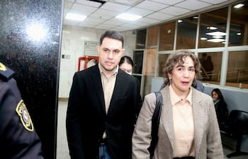Hombre con saco negro y camisa beige conversando con mujer de atuendo a cuadros marrón en un ambiente formal.