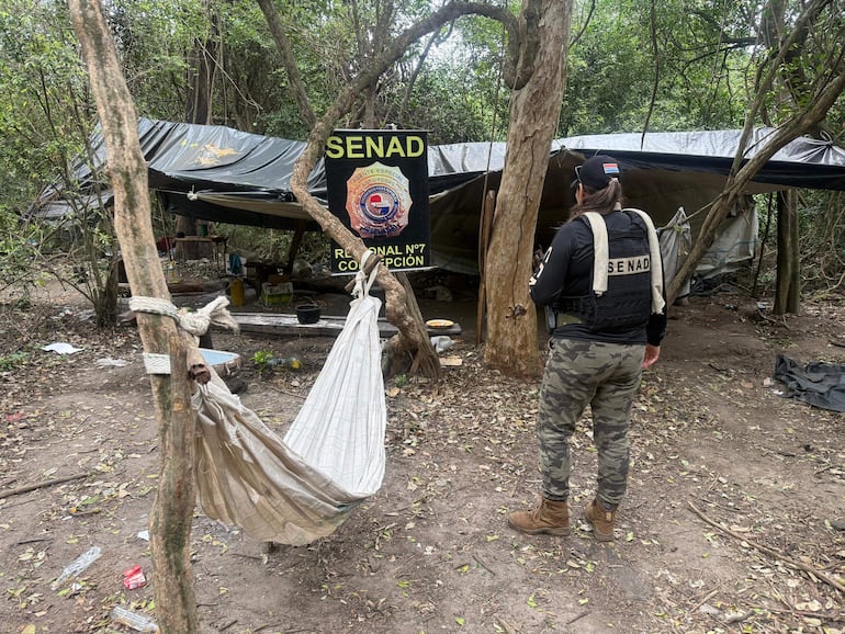 Mujer de espaldas en ropa táctica negra, con chaleco de la SENAD, en un campamento desordenado con toldo y hamaca.