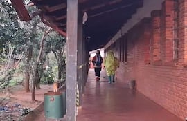 Dos bomberos, uno con traje amarillo impermeable y otro en chaqueta oscura, caminan en un pasillo de ladrillo rodeado de vegetación.