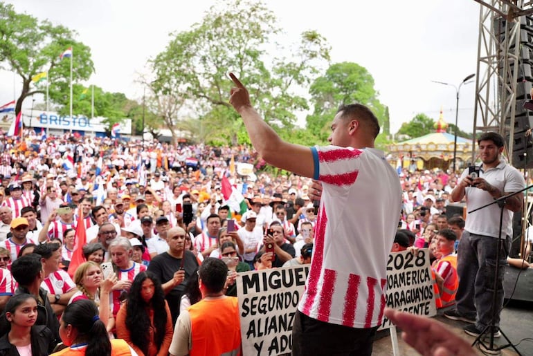 Hombre en camiseta de Club Olimpia habla a emocionado público con banderas paraguayas y carteles, en un ambiente festivo al aire libre.