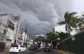 Calle urbana en Asunción con nubes oscuras, un automóvil blanco estacionado y un vehículo verde en movimiento.