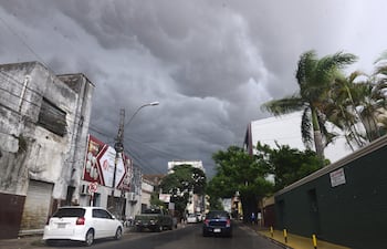 Calle urbana en Asunción con nubes oscuras, un automóvil blanco estacionado y un vehículo verde en movimiento.