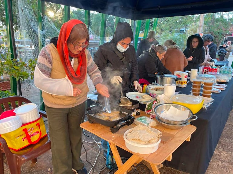 Grupo de mujeres cocinan en mesa larga, con alimentos y utensilios. Ambiente de feria gastronómica en día frío.