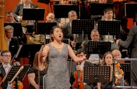 Cantante de ópera en vestido plateado interpreta apasionadamente al público, rodeada de músicos de orquesta en el escenario del teatro.