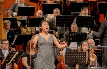 Cantante de ópera en vestido plateado interpreta apasionadamente al público, rodeada de músicos de orquesta en el escenario del teatro.