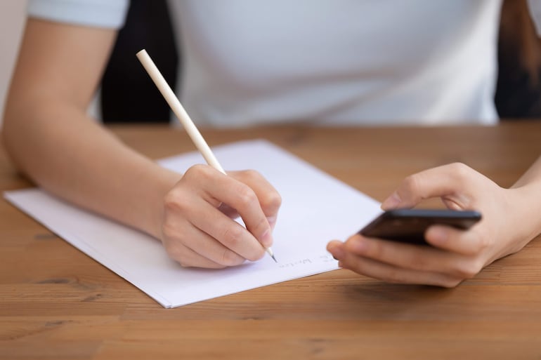 Mano escribiendo con lápiz en papel, sosteniendo un teléfono móvil con la otra mano en un entorno tranquilo.