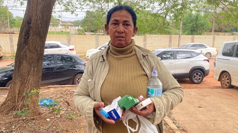 Mujer de mediana edad con camiseta beige y chaqueta, sostiene productos de salud y agua. Fondo con vehículos y vegetación.