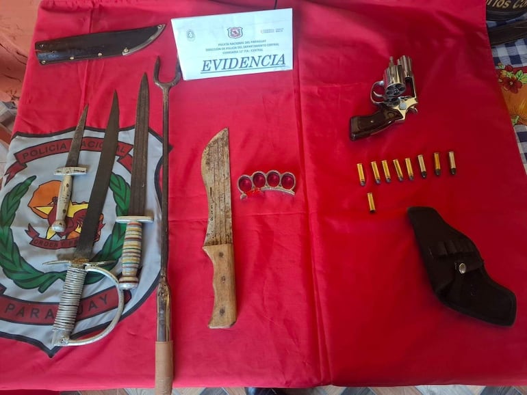 Conjunto de armas, incluido un cuchillo y una pistola, sobre un paño rojo con un escudo policial visible en el fondo.