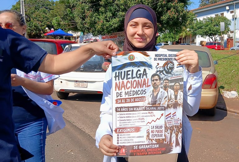 Mujer con blusa blanca y pañuelo en la cabeza sostiene un cartel en una manifestación, acompañada por otra mujer médico en uniforme.