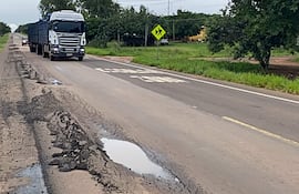 Camión cargado avanza por carretera con baches y charcos, rodeado de vegetación verde y señalización vial.