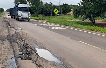 Camión cargado avanza por carretera con baches y charcos, rodeado de vegetación verde y señalización vial.
