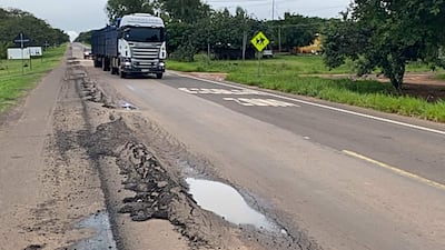 Camión cargado avanza por carretera con baches y charcos, rodeado de vegetación verde y señalización vial.