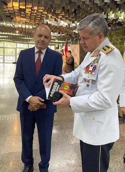 Pedro Lesme en traje oscuro escucha a Carlos Benítez en uniforme blanco, sosteniendo un libro titulado 'CÓDIGO' en ambiente institucional.