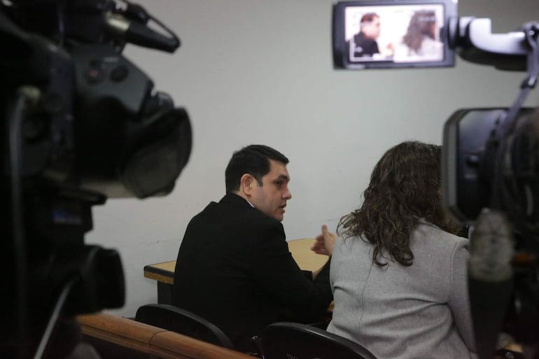 Hernán Rivas, en saco negro, habla en una mesa junto a una mujer con chaqueta gris, rodeados de cámaras en sala de prensa.