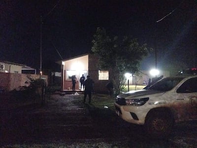 Tres personas vestidas de oscuro en la entrada de una casa iluminada, con una camioneta blanca en primer plano.