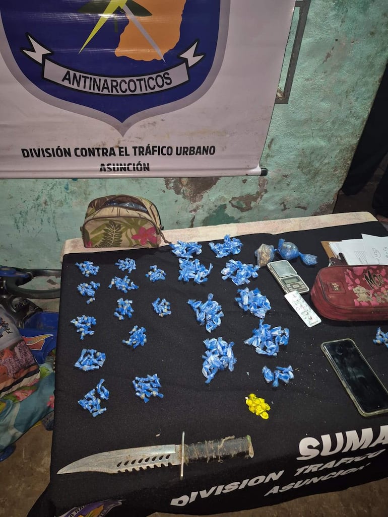 Mesa cubierta con mantel negro, envoltorios azules y amarillos, cuchillo, balanza y teléfono móvil en ambiente policial.