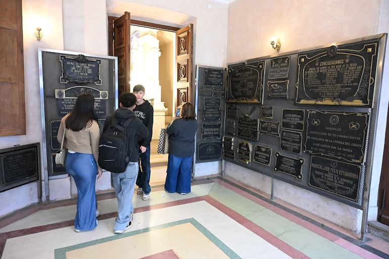 Una mujer de espaldas y tres jóvenes conversan en un museo con placas conmemorativas, en un ambiente solemne.