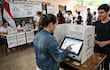 Joven de espaldas interactúa con máquina de votación, mientras otro sostiene su documento de identidad en un ambiente electoral animado.