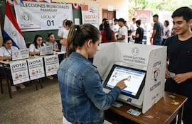 Joven de espaldas interactúa con máquina de votación, mientras otro sostiene su documento de identidad en un ambiente electoral animado.