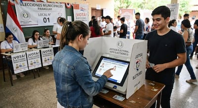 Joven de espaldas interactúa con máquina de votación, mientras otro sostiene su documento de identidad en un ambiente electoral animado.