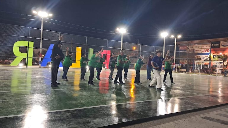Grupo de adultos mayores uniformados con camisetas verdes, levantando los brazos en coordinación bajo una cancha iluminada.
