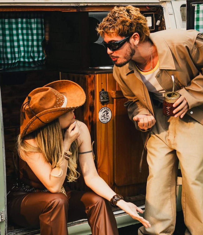 Mujer con sombrero y hombre con gafas oscuras conversan al lado de una furgoneta vintage, con una tela a cuadros verdes de fondo.