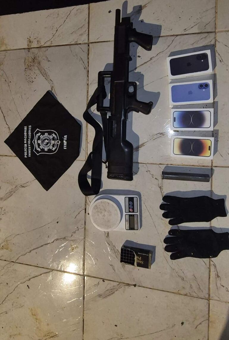 Escena interior con un arma larga, teléfonos en caja y guantes negros pertenecientes a una investigación policial.