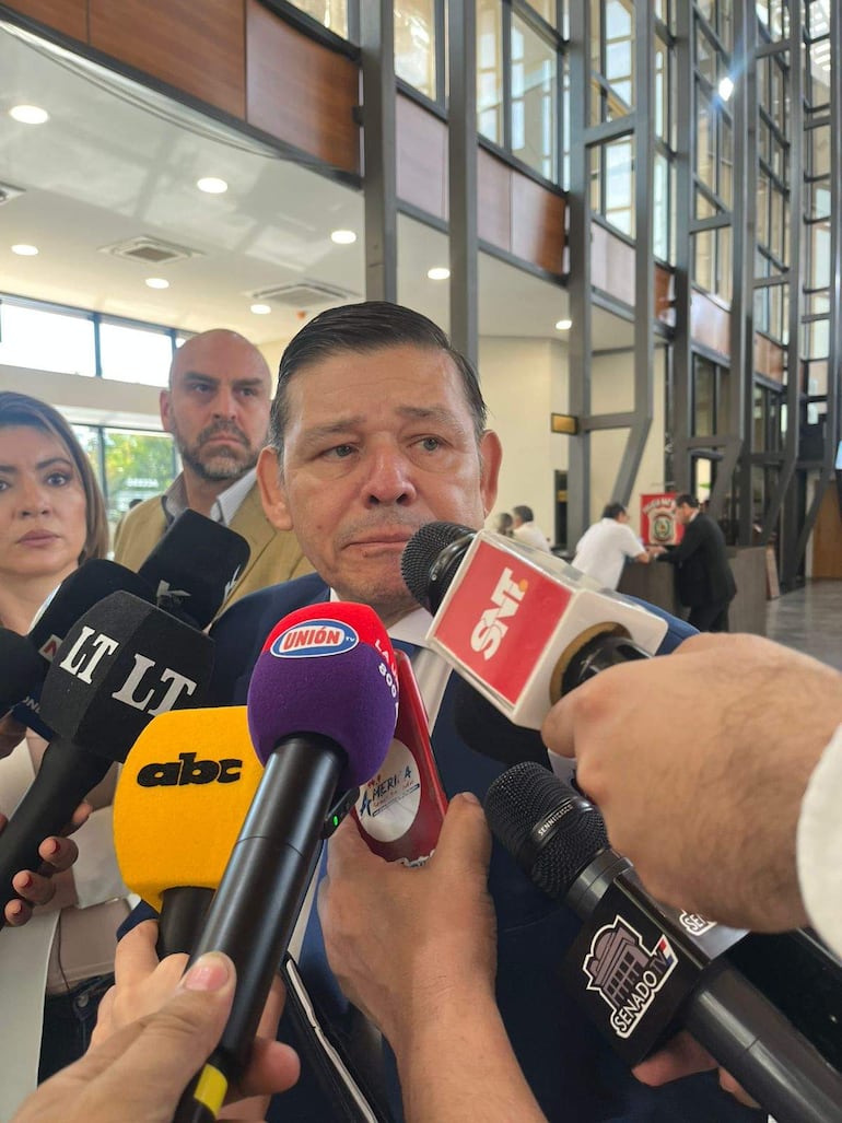 Hombre en traje oscuro muestra preocupación mientras dos personas lo acompañan en audiencia pública, rodeados de micrófonos.