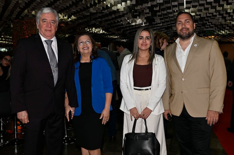Cuatro personas posan en ambiente elegante: hombre de traje oscuro, mujer con blusa negra y saco azul, otra mujer en conjunto blanco, y hombre de saco claro.