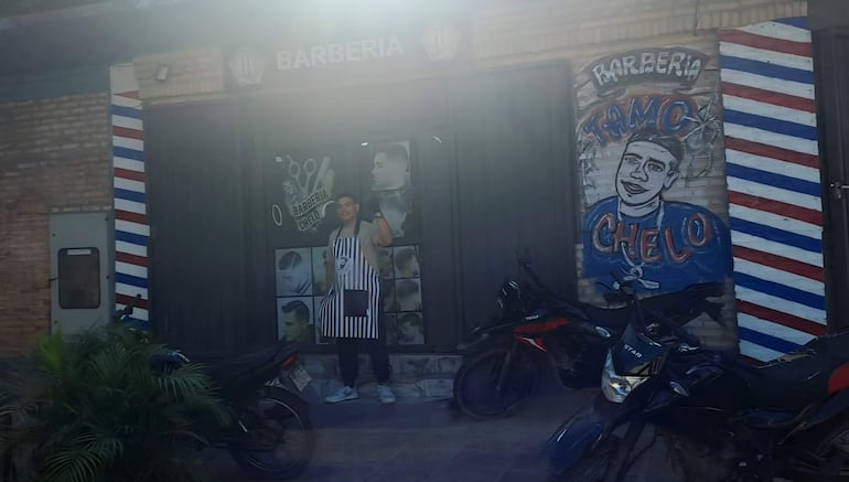 Hombre en delantal a rayas sonríe y saluda desde la entrada de Barbería Chelo, rodeado de motocicletas estacionadas.