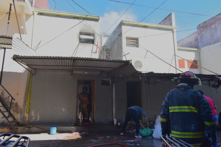 Bombero con traje y casco controlando un incendio, mientras una persona agachada recoge objetos en un saco en un entorno urbano.