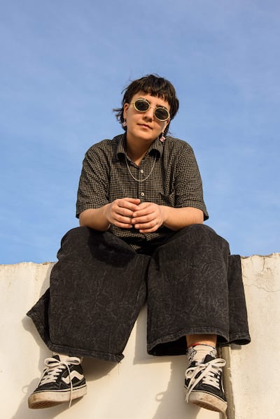 Sol Gómez con gafas de sol, camiseta de cuadros oscuros y pantalones negros, sentada con las piernas cruzadas bajo un cielo despejado.