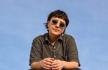 Sol Gómez con gafas de sol, camiseta de cuadros oscuros y pantalones negros, sentada con las piernas cruzadas bajo un cielo despejado.