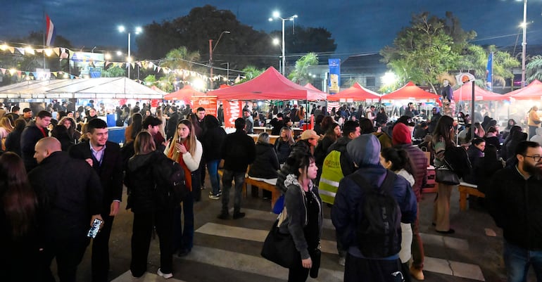 Multitud de personas en bazar nocturno, conversando y disfrutando de la comida en un ambiente festivo con luces y carpas rojas.