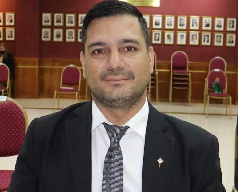 Hombre con cabello oscuro y barba, vestido con traje negro y corbata gris, en un entorno institucional con sillas rojas apiladas.