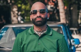 Arcángel de pie frente a un auto clásico, viste camiseta polo verde con detalles blancos y gafas de sol, en un entorno con vegetación.