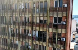 Varias personas asomadas en diferentes pisos de un edificio, una sostiene una bandera, en un ambiente urbano.