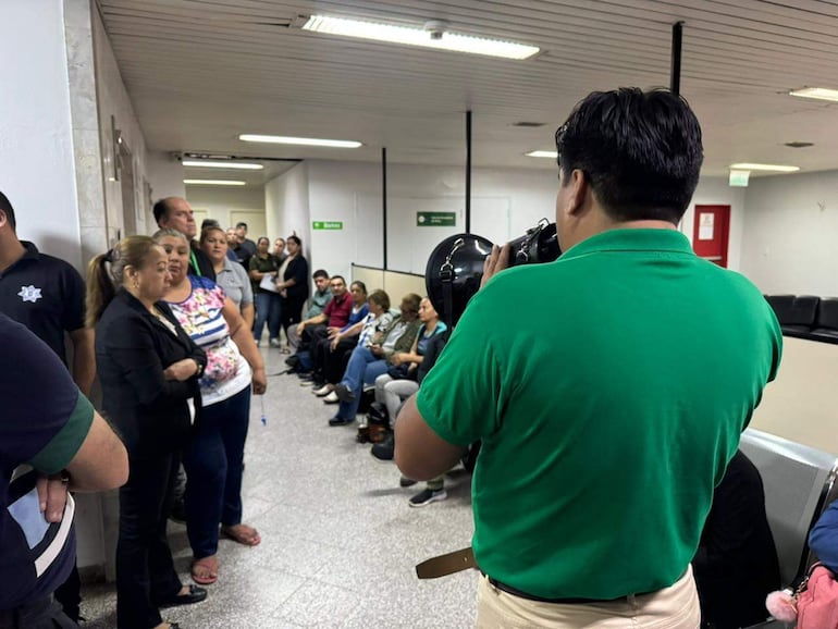 Grupo de personas en un pasillo, mujer con cabello castaño atenta a un hombre de espaldas con micrófono, ambiente interior.