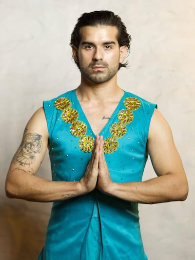 Hombre en vestido aqua con detalles dorados, de pie en posición de oración, con fondo neutro y expresión seria.