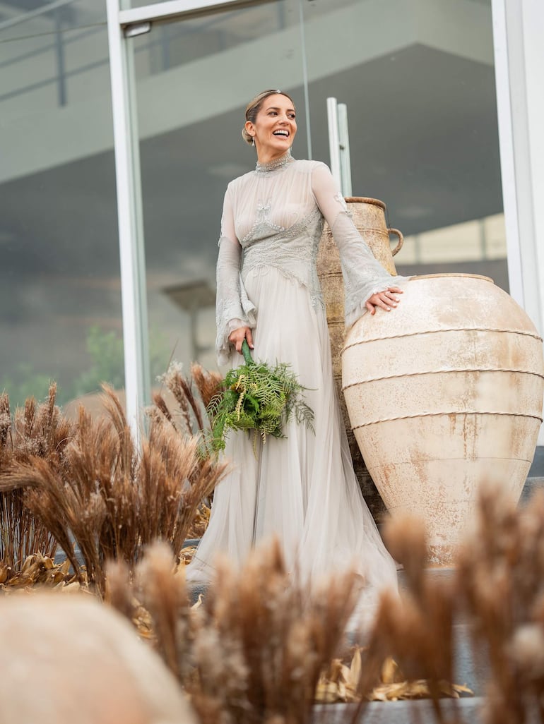 Emma Viedma sonríe en un vestido de novia claro, sosteniendo un ramo de hojas verdes, en un ambiente decorado con plantas.