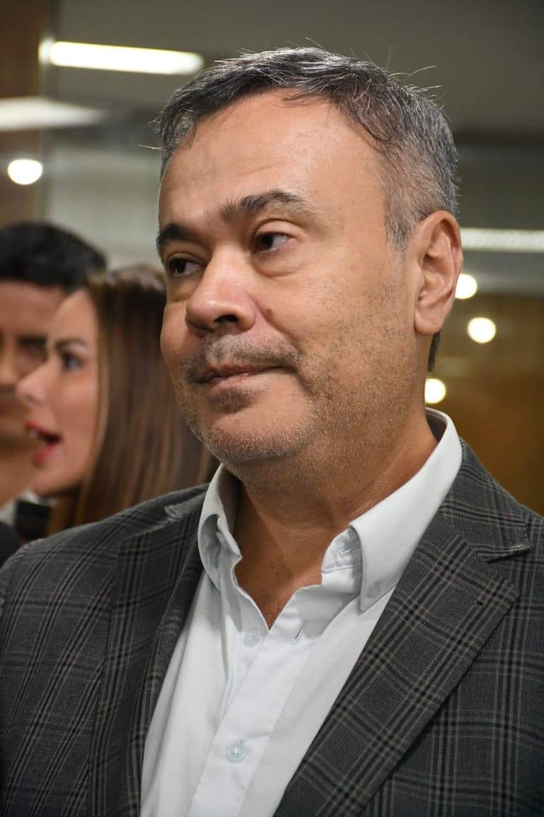 Humberto Blasco con expresión seria, lleva saco a cuadros; mujer a su izquierda habla, y otro hombre lo observa.