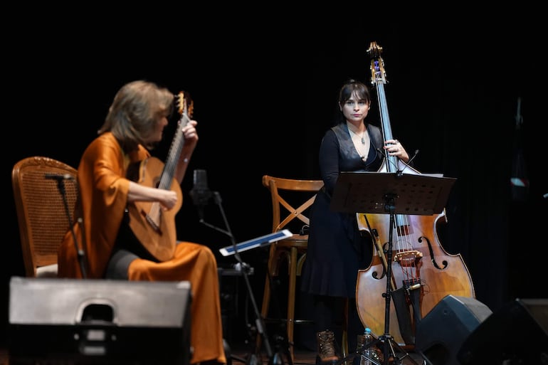 Berta Rojas con cabello oscuro y vestido oscuro, tocando el contrabajo, mientras su compañera rubia en atuendo naranja toca la guitarra.