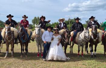 Johana Denis en vestido blanco junto a Alberto Roa, rodeados de seis hombres a caballo, todos sonrientes en un ambiente festivo.