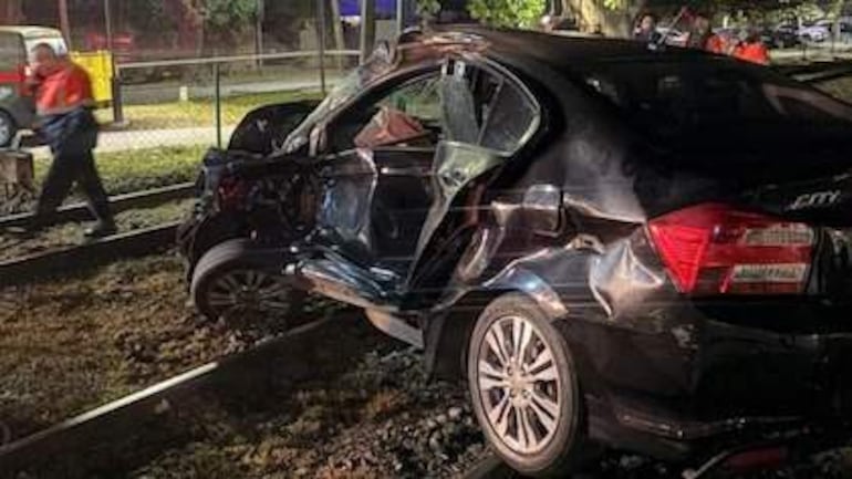 Automóvil destrozado en rieles de tren. Puerta del conductor abierta, luces intermitentes encendidas en un ambiente exterior.